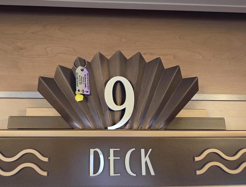 Puede incluir: Un letrero de metal marr&oacute;n con un dise&ntilde;o de abanico decorativo y el n&uacute;mero 9 en blanco. El letrero tambi&eacute;n dice "Deck" en letras blancas.
