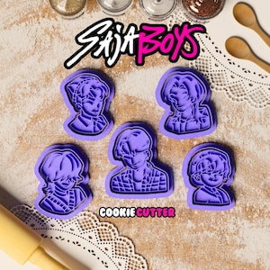 May include: A collection of five purple plastic cookie cutters in the shape of stylized male figures. The word "SAJA BOYS" is written in white and pink above the cutters, and "COOKIE CUTTER" is written in pink below.