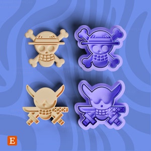 Op de afbeelding: Twee sets koekjesuitstekers, een paarse en een beige, met het iconische schedel- en gekruiste beenderen symbool uit de populaire anime-serie One Piece. De schedel draagt ​​een strohoed en een sjaal om zijn nek. De gekruiste beenderen zijn zwaarden.