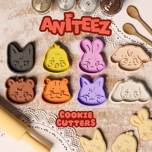 May include: A set of 10 colorful plastic cookie cutters featuring various animal designs, including a cat, a chick, a bunny, a dog, a bear, a tiger, a fox, a puppy, and two bunnies. The cutters are arranged on a wooden surface with a white doily and the text "AWITEEZ COOKIE CUTTERS".
