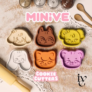 May include: A set of nine cookie cutters in various shapes, including a fox, a dog, a cat, and a bear. The cutters are made of a brown, pink, orange, and white plastic. The cutters are arranged on a white lace tablecloth with the words "MINIVE COOKIE CUTTERS" printed in pink and white. The background is a light brown surface.