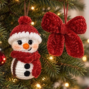 May include: A crochet snowman ornament wearing a red hat and scarf with black buttons, alongside a separate red crochet bow ornament. Both items feature soft, textured yarn and are suspended by thin brown and red strings for hanging.