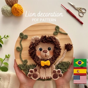 Könnte beinhalten: Gehäkelte Löwen-Dekoration auf einer Holzplatte. Der Löwe hat eine braune Mähne, eine gelbe Fliege und ist von gehäkeltem grünen Laub umgeben. Der Text "Lion decoration PDF PATTERN" ist sichtbar.