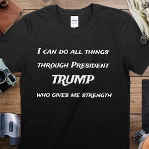Puede incluir: Camiseta negra con texto blanco: "I CAN DO ALL THINGS THROUGH PRESIDENT TRUMP WHO GIVES ME STRENGTH." La camiseta se muestra sobre una superficie de madera, junto con una cámara, unos prismáticos y otros accesorios.