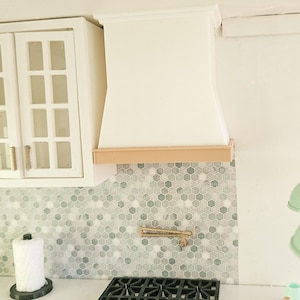 May include: Miniature kitchen scene with a white range hood, hexagonal tile backsplash, and a black gas cooktop. Two mint green pendant lights hang above a white countertop. A paper towel holder and a mint green cabinet are also visible.