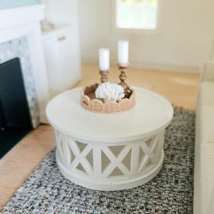 May include: A miniature white round coffee table with a decorative lattice design. A woven tray holds two white candles in wooden holders and a white coral decoration. The table sits on a blue and gray woven rug.