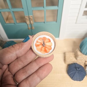 Könnte beinhalten: Ein runder Miniatur-Holzrahmen mit einem weißen Einsatz, der eine Illustration eines orangefarbenen Kürbisses im Aquarellstil zeigt. Der Rahmen verfügt über eine strukturierte, seilartige Randverzierung und ist ein kleines Deko-Objekt.
