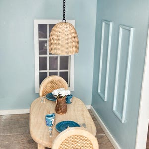 May include: Miniature dining room scene with a light wood table and two woven chairs. A woven pendant light hangs above the table, which is set with blue plates, cups, and a vase of white flowers. The walls are painted light blue.
