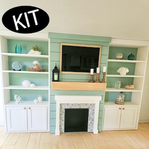 Può includere: Un set da soggiorno in miniatura per casa delle bambole con camino, librerie e TV. Il camino ha un caminetto bianco e una cornice di piastrelle grigie. Le librerie sono bianche con schienale blu e sono piene di oggetti decorativi. La parola "KIT" è in un ovale nero.