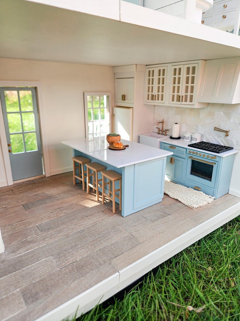Puede incluir: Una cocina de casa de mu&ntilde;ecas con una isla azul claro, gabinetes blancos y una encimera blanca. La isla tiene tres taburetes de bar y una peque&ntilde;a planta en maceta. La cocina tiene un salpicadero de azulejos blancos y una estufa de acero inoxidable.