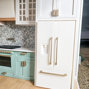 May include: A miniature kitchen model with a white fridge, gold handles, and a water dispenser. Mint green cabinets with gold hardware sit beside a black hob and white extractor fan. The kitchen has a light wood floor and a mosaic tile splashback.