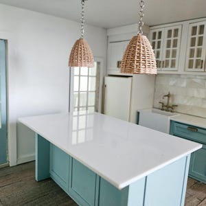 Könnte beinhalten: Miniatur-Kücheninsel mit weißer Arbeitsplatte und hellblauem Sockel. Zwei gewebte Pendelleuchten hängen über der Insel. Die Küche verfügt über weiße Schränke und einen Kühlschrank, wodurch ein heller und luftiger Raum entsteht.