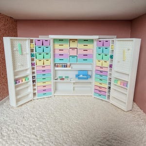 Puede incluir: Un armario de habitación artesanal blanco en miniatura con cajones y estantes de colores pastel. Las puertas del armario se abren para revelar espacio de almacenamiento para materiales de artesanía, incluidos cajones, estantes y tableros perforados. La habitación está decorada con una pared rosa y una alfombra blanca.