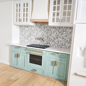 May include: Miniature kitchen with light blue cabinets, white countertops, and a mosaic tile backsplash. Features a black stovetop, oven, and gold-toned hardware. Includes white upper cabinets and a range hood. The floor is made of light-colored wood.