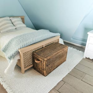 Puede incluir: Una escena de dormitorio en miniatura con una estructura de cama de madera clara y un cabecero tejido. Un cofre de almacenamiento marrón tejido se encuentra al pie de la cama. La cama está hecha con ropa de cama blanca y una manta azul. Hay una alfombra blanca en el suelo.