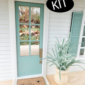 May include: A miniature dollhouse door with a light blue door and a welcome mat that says "Hello Sunshine" with a sun graphic. The door is framed in white and has a gold door handle. A potted plant with green leaves is to the right of the door.