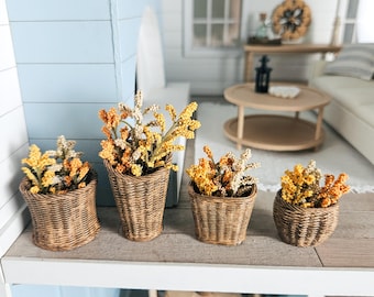 May include: Four miniature woven wicker baskets, each filled with dried yellow and orange floral arrangements. The baskets are arranged on a white shelf, with a light blue wall in the background. The scene evokes a coastal or farmhouse aesthetic.