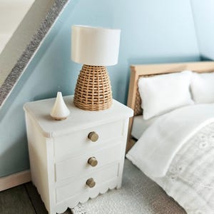 May include: A miniature bedroom scene featuring a white nightstand with three drawers and gold knobs. A woven lamp with a white shade sits atop the nightstand, next to a small white decorative object. A bed with white bedding is in the background.