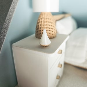 Könnte beinhalten: Ein weißer Nachttisch mit drei Schubladen und goldfarbenen Griffen. Auf der Oberseite steht eine gewebte Lampe mit weißem Lampenschirm sowie ein weißes, kegelförmiges Dekorationsobjekt. Der Hintergrund ist eine hellblaue Wand.