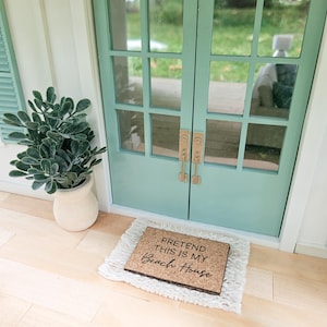 May include: A miniature beach house scene featuring a light teal double door with glass panes and brass handles. A doormat with the text "PRETEND THIS IS MY Beach House" sits on a white rug. A potted plant is to the left.