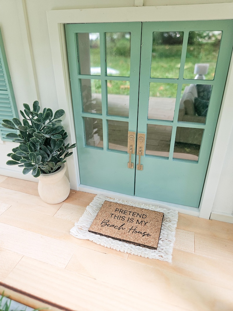 May include: A miniature beach house scene featuring a light teal double door with glass panes and brass handles. A doormat with the text "PRETEND THIS IS MY Beach House" sits on a white rug. A potted plant is to the left.