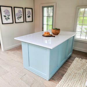 Puede incluir: Una isla de cocina azul claro con una encimera blanca. Un peque&ntilde;o pastel y una planta en maceta est&aacute;n en la encimera. La isla est&aacute; en una habitaci&oacute;n con paredes blancas y pisos de madera.