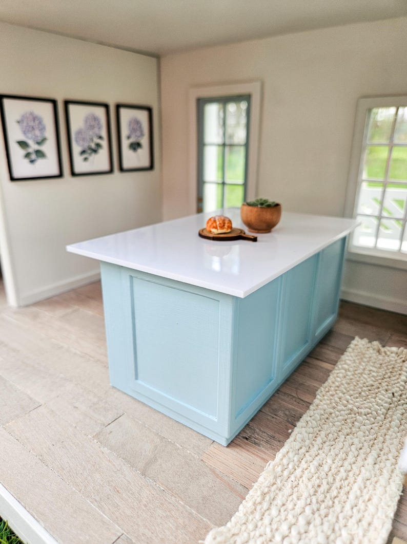 Puede incluir: Una isla de cocina azul claro con una encimera blanca. Un peque&ntilde;o pastel y una planta en maceta est&aacute;n en la encimera. La isla est&aacute; en una habitaci&oacute;n con paredes blancas y pisos de madera.