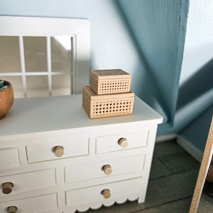 May include: A miniature white dresser with gold knobs and a scalloped edge. Two small woven boxes are stacked on top. The background features a light blue wall and a white window frame.