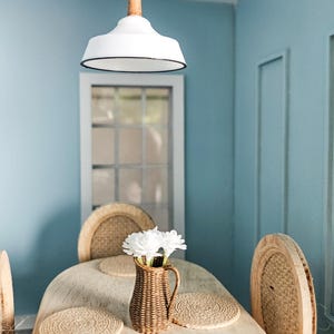 Può includere: Una scena di sala da pranzo in miniatura con una parete azzurra e una lampada a sospensione bianca. Un tavolo di legno è apparecchiato con sottopiatti intrecciati, sedie e un vaso di vimini con fiori bianchi. La stanza ha un'estetica rustica e accogliente.
