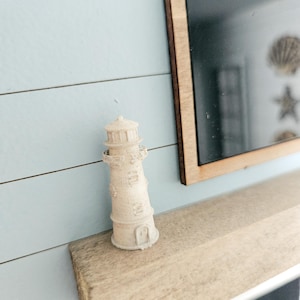 May include: A small, off-white lighthouse figurine with detailed architectural features, standing on a light-colored wooden shelf. The background features a light blue wall with horizontal paneling and a framed artwork with nautical-themed decorations.