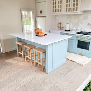 May include: A dollhouse kitchen island with a white countertop and light blue cabinets. The island has four wooden bar stools and a small plate of pastries on the countertop.