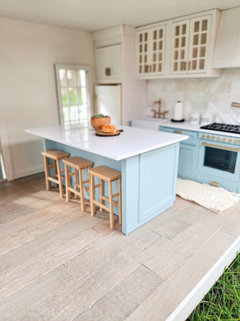 May include: A dollhouse kitchen island with a white countertop and light blue cabinets. The island has four wooden bar stools and a small plate of pastries on the countertop.