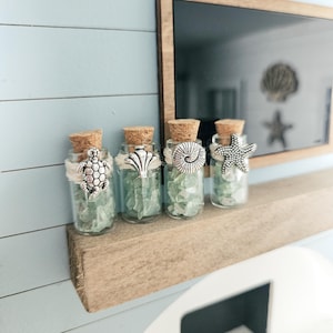 Puede incluir: Cuatro pequeñas botellas de vidrio con tapones de corcho, cada una con vidrio de mar y un dije decorativo. Los dijes incluyen una tortuga, una concha, un nautilus y una estrella de mar. Tema costero.