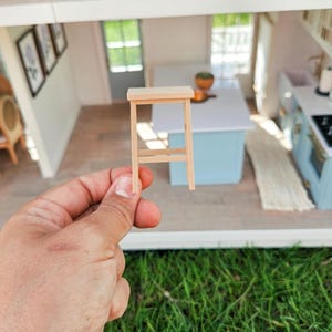 May include: A miniature wooden stool with a light brown finish. The stool is being held in a hand and is in focus, while the background is a blurry image of a dollhouse interior.