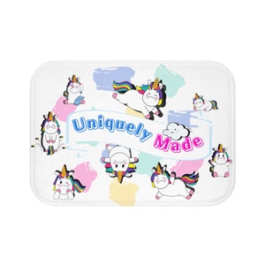 May include: A white bath mat with a colorful design featuring multiple cartoon unicorns in various poses. The text "Uniquely Made" is written in a blue banner with a pink cloud above it.