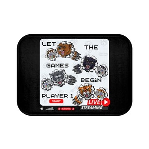 May include: Black doormat with a white background featuring a cartoon illustration of four animals, a bear, a wolf, a panther, and a tiger, each holding a video game controller. The text "Let the games begin" is written in a pixelated font. The text "Player 1" is written below the animals. The text "Live Streaming" is written below the text "Player 1".