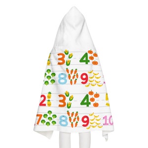 Op de afbeelding: Witte badhanddoek met capuchon met kleurrijke cijfers en cartoon groenten. Het ontwerp bevat broccoli, wortelen, maïs, bananen en pompoenen. De cijfers zijn in verschillende kleuren en gerangschikt in rijen.
