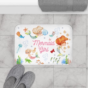 Könnte beinhalten: Weiße Badematte mit Meerjungfrauen-Motiv. Die Matte zeigt zwei Meerjungfrauen mit rosa Schwänzen, einen Seestern, ein Seepferdchen und die Worte "Mermaid Girl" in rosa Schrift.