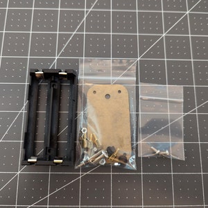 May include: A black battery holder with two compartments, a small bag of brass screws and standoffs, and a brown cardboard circuit board.