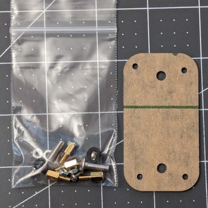 May include: A clear plastic bag containing various small metal hardware components, including screws, nuts, and bolts, sits next to a rectangular, tan-colored acrylic piece with rounded corners and several drilled holes. A green line is visible.