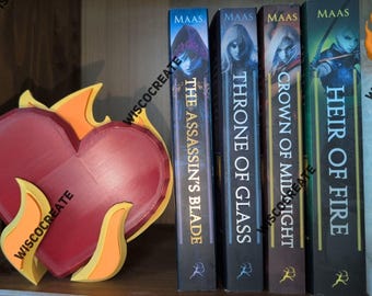 May include: A red heart-shaped decor with yellow and orange flames, next to four books with the titles "The Assassin's Blade", "Throne of Glass", "Crown of Midnight", and "Heir of Fire". The books have a dark background.
