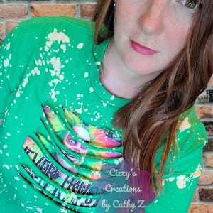 Könnte beinhalten: Ein hellgrünes Kurzarm-T-Shirt mit gebleichtem Spritzmuster. Das Shirt zeigt einen Grafikdruck einer Figur mit buntem Haar und den Text "NEVER TRUST THE LIVING", der teilweise durch horizontale Stoffschlitze verdeckt wird.