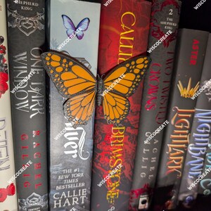Könnte beinhalten: Eine Sammlung von Hardcover-Büchern mit einer Monarchfalter-Dekoration. Die Bücher haben Titel in Rot, Weiß und Blau, mit einer Vielzahl von Schriftarten. Der Schmetterling ist orange und schwarz, mit komplizierten Flügelmustern. Die Bücher sind vertikal angeordnet.