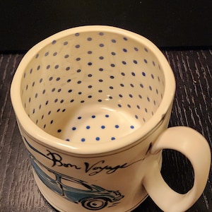 K&ouml;nnte beinhalten: Cremefarbene Keramik-Tasse mit blauen Tupfen im Inneren. Die Tasse zeigt eine blaue Auto-Illustration und die Worte "Bon Voyage" in Schreibschrift. Der Griff ist cremefarben.