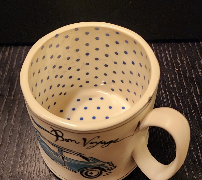 K&ouml;nnte beinhalten: Cremefarbene Keramik-Tasse mit blauen Tupfen im Inneren. Die Tasse zeigt eine blaue Auto-Illustration und die Worte "Bon Voyage" in Schreibschrift. Der Griff ist cremefarben.