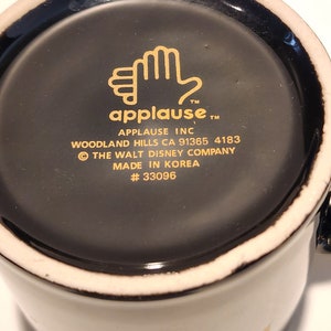 May include: Black ceramic mug with a gold rim and a gold design on the bottom. The design includes a hand with the word "applause" and the text "Applause Inc. Woodland Hills CA 91365 4183 &copy; The Walt Disney Company Made in Korea #33096"