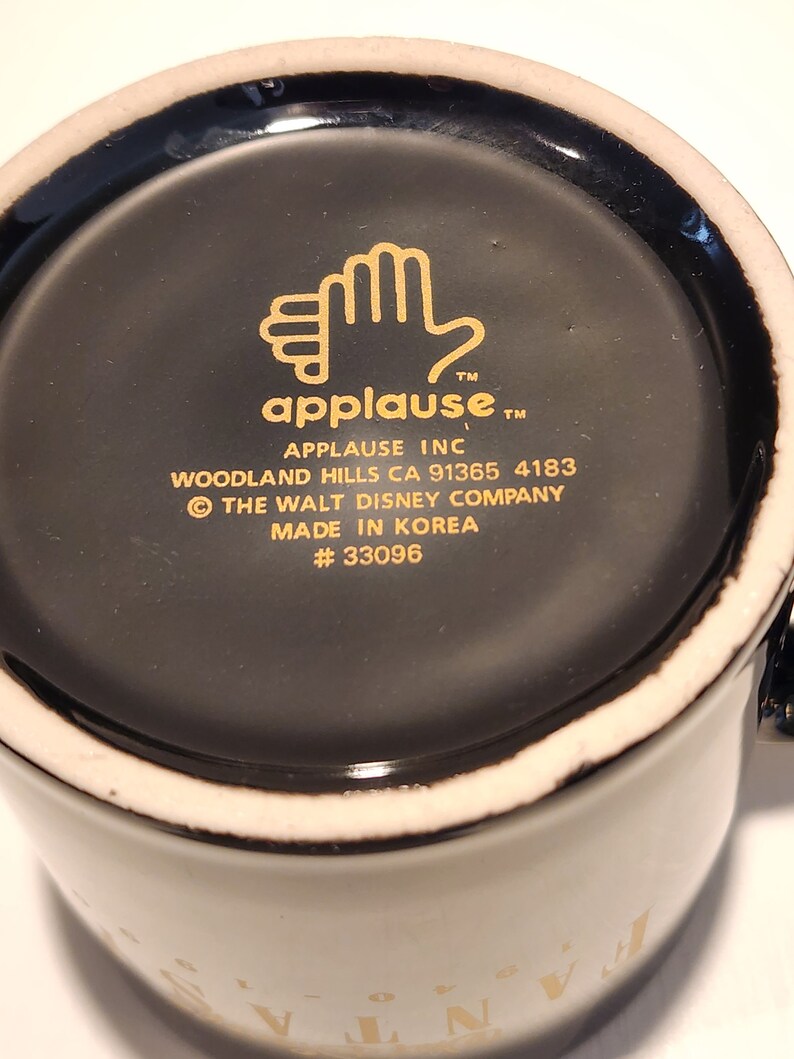 May include: Black ceramic mug with a gold rim and a gold design on the bottom. The design includes a hand with the word "applause" and the text "Applause Inc. Woodland Hills CA 91365 4183 &copy; The Walt Disney Company Made in Korea #33096"