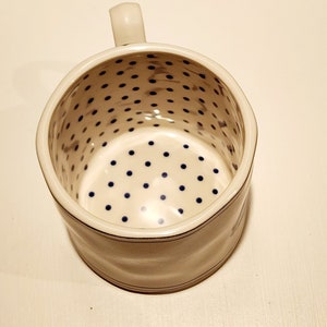 K&ouml;nnte beinhalten: Eine wei&szlig;e Rasierkelle aus Keramik mit Griff. Der Innenraum weist ein Muster aus blauen Punkten auf. Das &Auml;u&szlig;ere hat ein schlichtes Design mit einer d&uuml;nnen blauen Linie in der N&auml;he der Ober- und Unterseite.