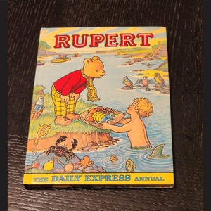 Puede incluir: Una portada de libro vintage con Rupert el Oso, un personaje de dibujos animados, con el t&iacute;tulo "Rupert The Daily Express Annual". La portada est&aacute; ilustrada con Rupert el Oso con un atuendo rojo y amarillo, sosteniendo una cesta de fruta, y un ni&ntilde;o nadando en el agua.