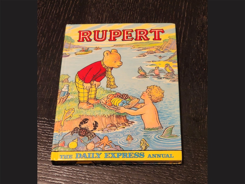 Puede incluir: Una portada de libro vintage con Rupert el Oso, un personaje de dibujos animados, con el t&iacute;tulo "Rupert The Daily Express Annual". La portada est&aacute; ilustrada con Rupert el Oso con un atuendo rojo y amarillo, sosteniendo una cesta de fruta, y un ni&ntilde;o nadando en el agua.